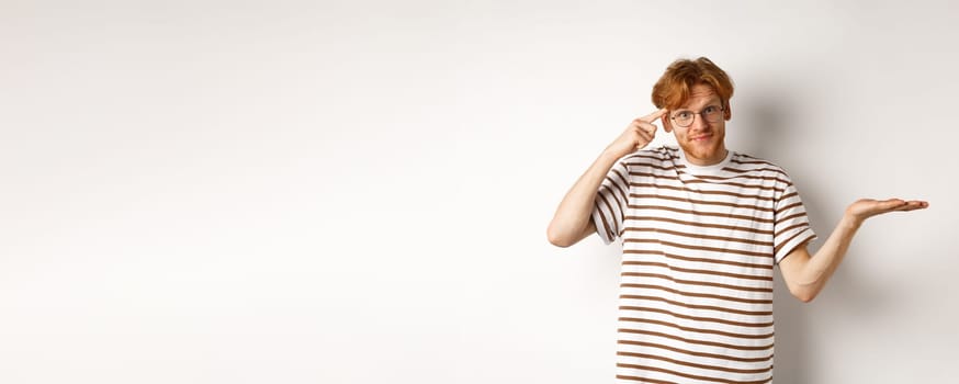 Are you stupid. Confused redhead guy in glasses shrugging, pointing at head and staring skeptical at camera, white background.