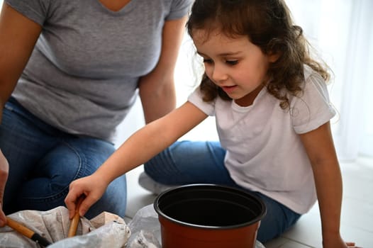 People, gardening and housework concept - little child using garden shovel, putting black soil in a plastic pot, helping her mom in gardening, repotting houseplants at springtime in the home veranda