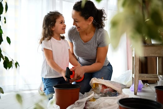 Adorable child, lovely baby girl smiles looking at her loving mother while helping her in floriculture, watering together planted seed at home. Earth Day. Environmental conservation. Save planet