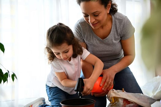 Adorable and caring lovely baby girl, wearing white t-shirt and casual denim, smiling, enjoying helping her mother to watering houseplants while repotting them in springtime. People. Gardening concept