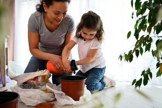 Lovely little girl watering potted soil while planting houseplants with her mom on home balcony. Woman from childhood teaches her daughter to take care of nature. Concept of environmental conservation