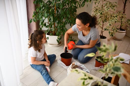 Happy loving mother watering back soil with plant seeds in plastic pots, taking care of houseplants and seedlings, smiling to cute child, enjoying gardening hobby in the home veranda in springtime