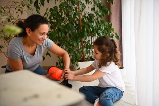 Beautiful African American woman loving mother teaching her adorable daughter to care for houseplants, smiling, enjoying together gardening in spring in a home veranda. People and environment concept