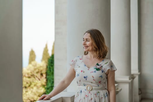 a beautiful mature woman in a long white dress stands on the balcony and looks into the distance.