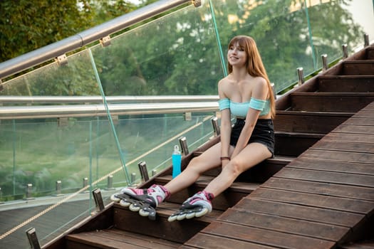 A long-haired brunette in a strapless top sits on the stairs. The girl is wearing shorts and roller skates. The woman is resting on a wooden staircase sitting by a glass railing. The treetops can be seen in the background.