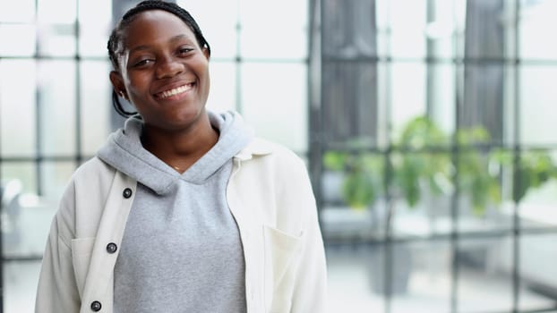 Smiling young woman standing and looking at the camera. She stands in a modern office against the background of a window.