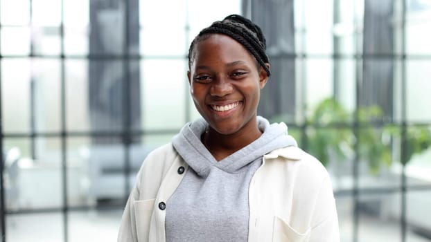 Smiling young woman standing and looking at the camera. She stands in a modern office against the background of a window.