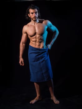 Portrait of naked handsome young man with languishing look covering crotch with a towel, against black background