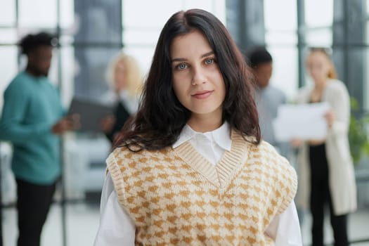 Portrait of cheerful young woman standing in office with coworkers talking in background.