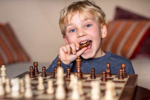 Small child 5 years old playing a game of chess on large chess board. Chess board on table in front of the boy , happily eating the lost piece