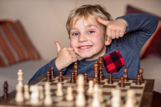Small child 5 years old playing a game of chess on large chess board. Chess board on table in front of the boy he is happy to win, showing thumbs up