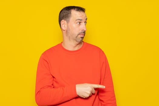 Bearded hispanic man wearing a red sweater pointing to the side with the index finger isolated on yellow studio background.