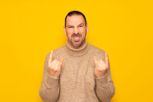 Bearded hispanic man wearing a beige turtleneck sweater making the rock gesture with aggressive and rebellious expression, isolated on yellow studio background