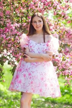 Beautiful smiling brunette girl in light pink dress, is standing near a pink blooming apple trees, in the spring in the garden. Looks to the side. Vertical. Copy space