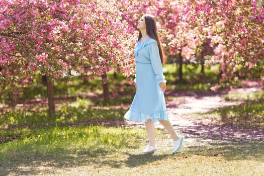 Happy brunette girl in light blue dress, with long hair is standing in a pink blooming garden, in sunny day. Copy space
