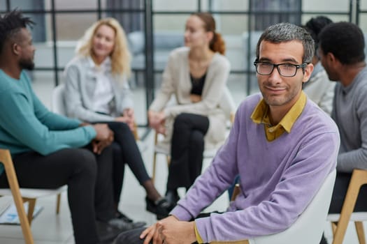 A happy young businessman, in a purple sweater, sits against the background of his colleagues in a modern office