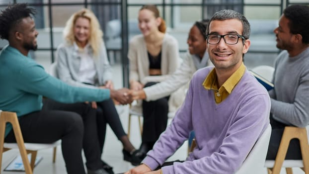 A happy young businessman, in a purple sweater, sits against the background of his colleagues in a modern office