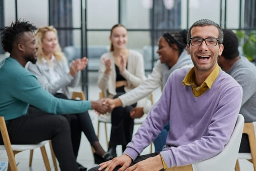 A happy young businessman, in a purple sweater, sits against the background of his colleagues in a modern office