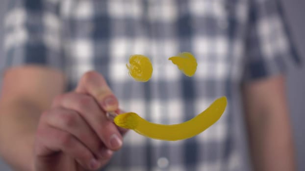 A young man draws a happy smile on the glass. A man in a plaid shirt draws with a brush and yellow paint. Close-up. 4k