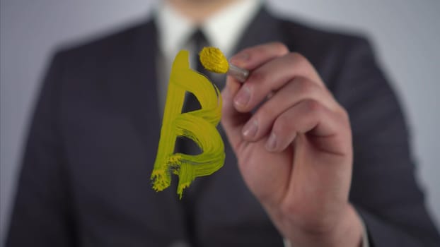 A young businessman draws a bitcoin symbol on a transparent glass. A man draws with yellow paint and a brush close-up. 4k