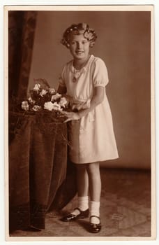 LIBEREC (REICHENBERG), THE CZECHOSLOVAK REPUBLIC - CIRCA 1930s: Vintage photo shows young girl with bouquet poses in a photography studio. Photo with dark sepia tint. Black & white studio portrait.