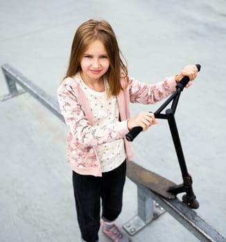 Preteen girl standing with scooter at street in the park and looking at camera. Cute child posing with eco vehicle outdoors