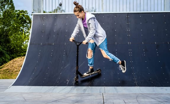 Pretty girl riding scooter on ramp in park. Cute teenager with eco vehicle outdoors