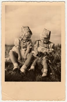 DRESDEN, GERMANY - CIRCA 1954: Vintage photo shows girls sit on grass. They wear traditional cap for girls. Antique black white photo.