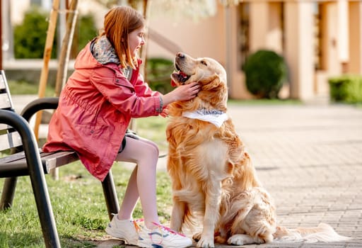 Girl petting golden retriever dog sitting on bench outdoors in sunny day in city. Female child kid and pet doggy labrador portrait