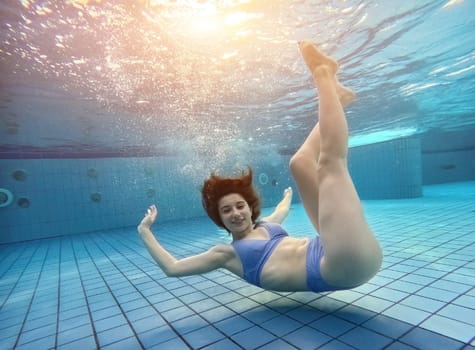 Beautiful teen girl swimming under water in blue pool and posing looking at camera. Pretty female teenager diving and having fun at summer