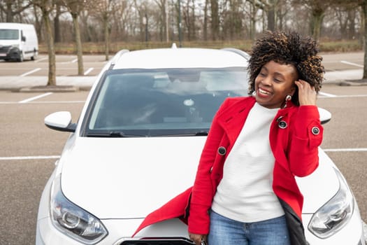 a cheerful african american woman stands next to the White car and laughing as he straightens his flowing hair, High quality photo