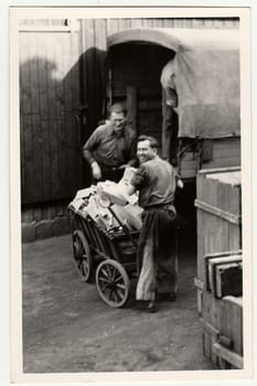 GERMANY - CIRCA 1950s: Vintage photo shows men upload the small boxes to lorry (truck). Antique black & white studio portrait.