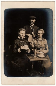 GERMANY - CIRCA 1910s: Vintage photo shows man and women pose with a cup, newspaper and pipe. Antique black & white studio portrait.