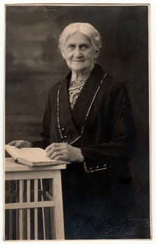 LIBEREC (REICHENBERG), THE CZECHOSLOVAK REPUBLIC - CIRCA 1920s: Vintage photo shows old woman with book poses in a photography studio. Photo with dark sepia tint. Black white studio portrait.