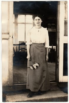 AUSTRIA-HUNGARY - 1915: Vintage photo shows woman stands in front of house and holds a lilac. Black white studio portrait.