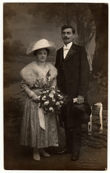 CHRASTAVA KRATZAU , THE CZECHOSLOVAK REPUBLIC - CIRCA 1920s: Vintage photo of newlyweds. Bride wears a furry shawl and white wide- brimmed hat with netting. Groom wears posh clothing, white bow-tie, gloves and black top hat. Black white antique studio portrait.