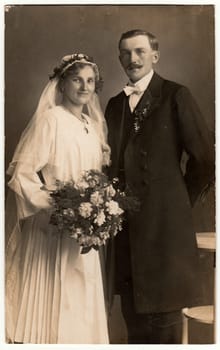 CHRASTAVA (KRATZAU), THE CZECHOSLOVAK REPUBLIC - CIRCA 1920s: Vintage photo of newlyweds. Bride wears veil and she holds wedding bouquet. Groom wears posh clothing, white bow-tie. Black white antique studio portrait.