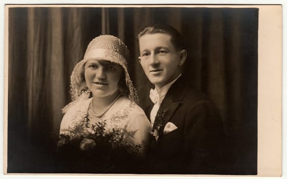 LIBEREC (REICHENBERG), THE CZECHOSLOVAK REPUBLIC - CIRCA 1920s: Vintage photo of newlyweds. Bride wears white hat with lace (bobbin). Groom wears posh clothing, white bow-tie. Black white antique studio portrait.