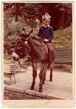 THE CZECHOSLOVAK SOCIALIST REPUBLIC - CIRCA 1970s: Vintage photo shows boy sits on donkey, he wears Indian headband and holds tomahawk. Colour photography.