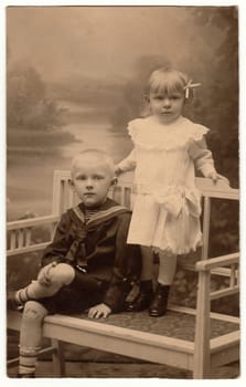 HRADEC KRALOVE, THE CZECHOSLOVAK REPUBLIC - AUGUST 11, 1930: Vintage photo shows two children (siblings). Boy wears sailor costum and girl wears white dress, laced collar and hair ribbon. They pose on historic white bench. Black white studio photography.