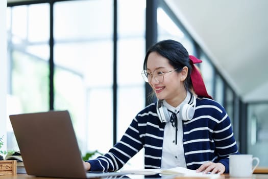 Portrait of a teenage Asian woman using a computer, wearing headphones and using a notebook to study online via video conferencing on a wooden desk in library.