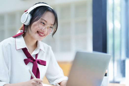 Portrait of a teenage Asian woman using a computer, wearing headphones and using a notebook to study online via video conferencing on a wooden desk in library.