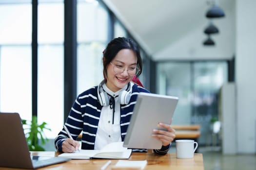 Portrait of a teenage Asian woman using a tablet, wearing headphones and using a notebook to study online via video conferencing on a wooden desk in library.