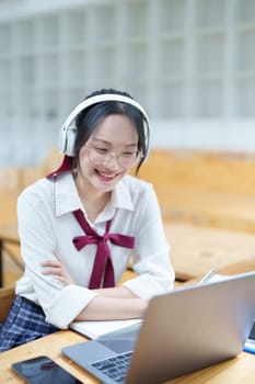 Portrait of a teenage Asian woman using a computer, wearing headphones and using a notebook to study online via video conferencing on a wooden desk in library.