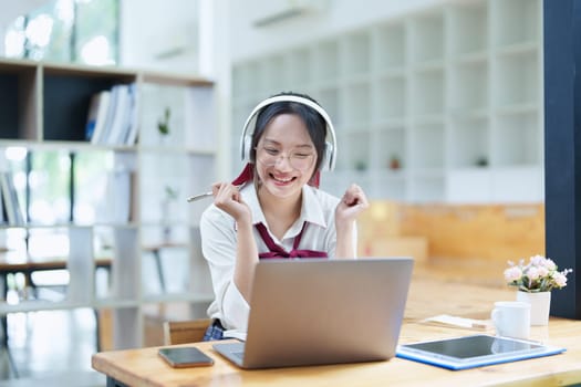 Portrait of a teenage Asian woman using a computer, wearing headphones and using a notebook to study online via video conferencing on a wooden desk in library.