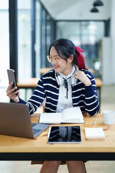 Portrait of a teenage Asian woman using a smartphone, wearing headphones and using a notebook to study online via video conferencing on a wooden desk in library.