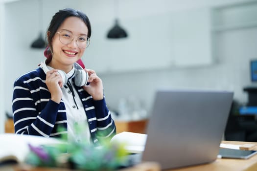 Portrait of a teenage Asian woman using a computer, wearing headphones and using a notebook to study online via video conferencing on a wooden desk in library.