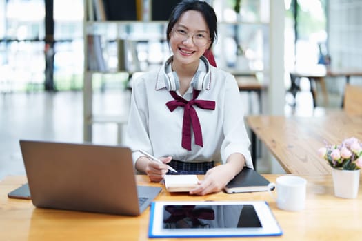 Portrait of a teenage Asian woman using a computer, wearing headphones and using a notebook to study online via video conferencing on a wooden desk in library.