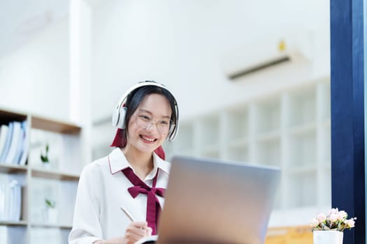 Portrait of a teenage Asian woman using a computer, wearing headphones and using a notebook to study online via video conferencing on a wooden desk in library.