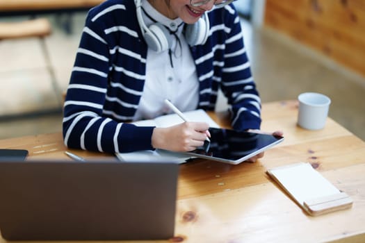 Portrait of a teenage Asian woman using a tablet, wearing headphones and using a notebook to study online via video conferencing on a wooden desk in library.